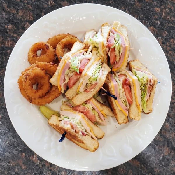 A white plate holds a club sandwich cut into quarters, onion rings, and a pickle spear.