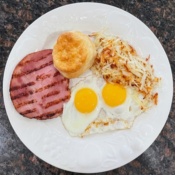 Breakfast plate with grilled ham, two fried eggs, hash browns, and a biscuit.
