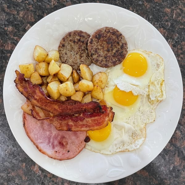 A full breakfast plate with fried eggs, sausage patties, bacon, ham, and roasted potatoes.